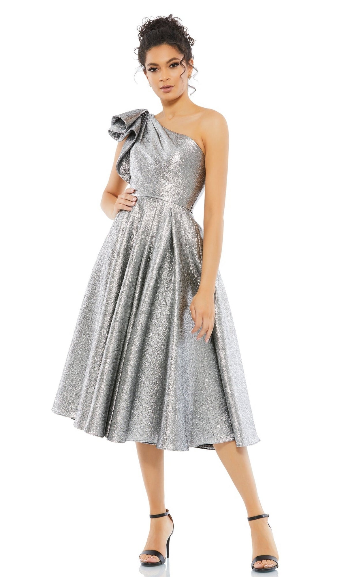 A woman stands against a white background in the Mac Duggal 67906 Metallic Short Wedding-Guest Dress, a knee-length one-shoulder style with bow detail, paired with black high-heeled sandals.